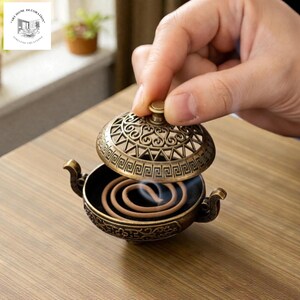 May include: An antique bronze incense burner with a decorative lid, featuring intricate patterns and a small handle. The burner holds a coiled incense stick, with smoke gently rising. The burner is on a wooden surface.