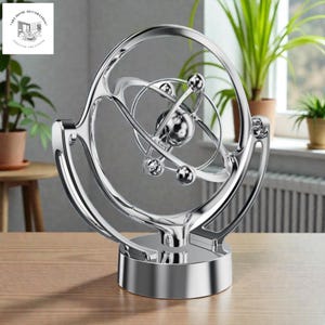 May include: A shiny silver kinetic sculpture with a spherical design. The sculpture has a central sphere with orbiting spheres and rings, mounted on a circular base. The sculpture is displayed on a wooden table.