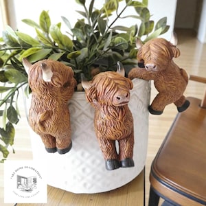 Puede incluir: Tres figuras de vacas de las Tierras Altas marrones con pelaje texturizado y pequeños cuernos están adheridas a una maceta blanca texturizada llena de follaje verde. Las figuras están en varias poses, agregando un toque caprichoso a la decoración del hogar.