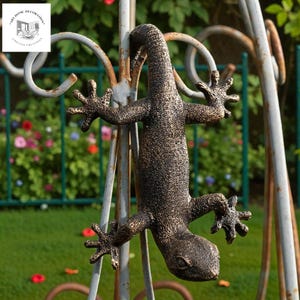May include: A bronze-colored lizard sculpture clinging to a decorative metal structure. The lizard has detailed skin texture and is positioned upside down. The background includes a green fence and foliage, suggesting an outdoor garden setting.