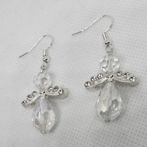 May include: A pair of clear crystal angel earrings. Each earring features a teardrop-shaped crystal body, a round crystal head, and silver-toned wings adorned with small, clear crystals. The earrings have silver-toned ear wires.