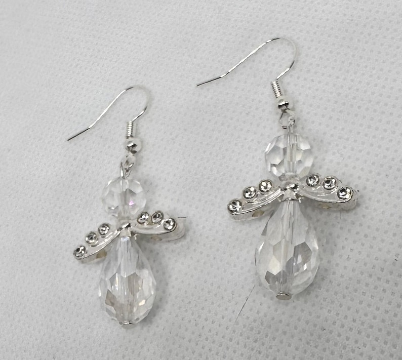 May include: A pair of clear crystal angel earrings. Each earring features a teardrop-shaped crystal body, a round crystal head, and silver-toned wings adorned with small, clear crystals. The earrings have silver-toned ear wires.