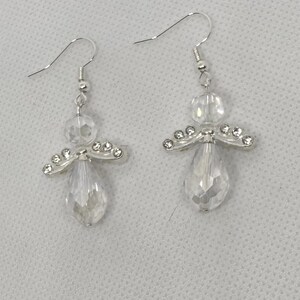 May include: A pair of clear crystal angel earrings. Each earring features a faceted teardrop-shaped crystal, a round crystal bead, and a silver-toned wing design with small clear crystals. The earrings have silver-colored fish hook ear wires.