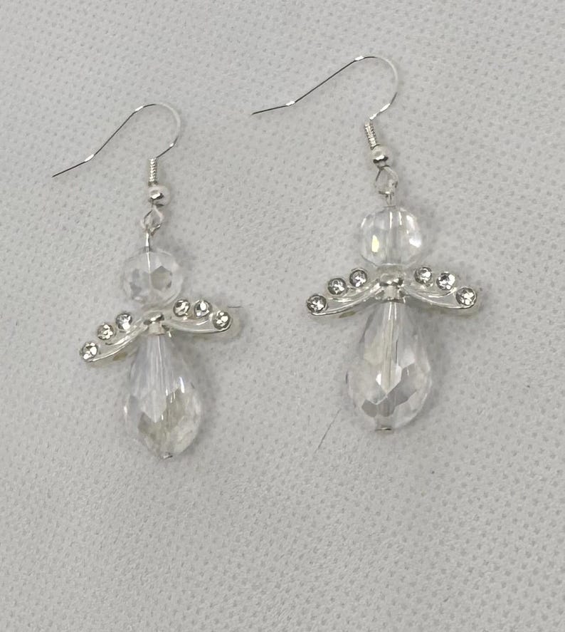 May include: A pair of clear crystal angel earrings. Each earring features a faceted teardrop-shaped crystal, a round crystal bead, and a silver-toned wing design with small clear crystals. The earrings have silver-colored fish hook ear wires.