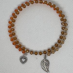 May include: A beaded bracelet with two strands of amber-colored beads. The bracelet features two silver charms: a heart and a leaf. The beads are strung on a flexible wire, creating a wrap-around design.