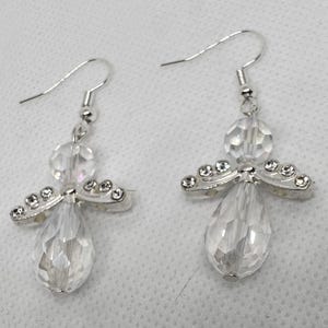 May include: A pair of clear crystal angel earrings. Each earring features a teardrop-shaped crystal body, a round crystal head, and silver-toned wings adorned with small, clear crystals. The earrings have silver-colored fish hook closures.
