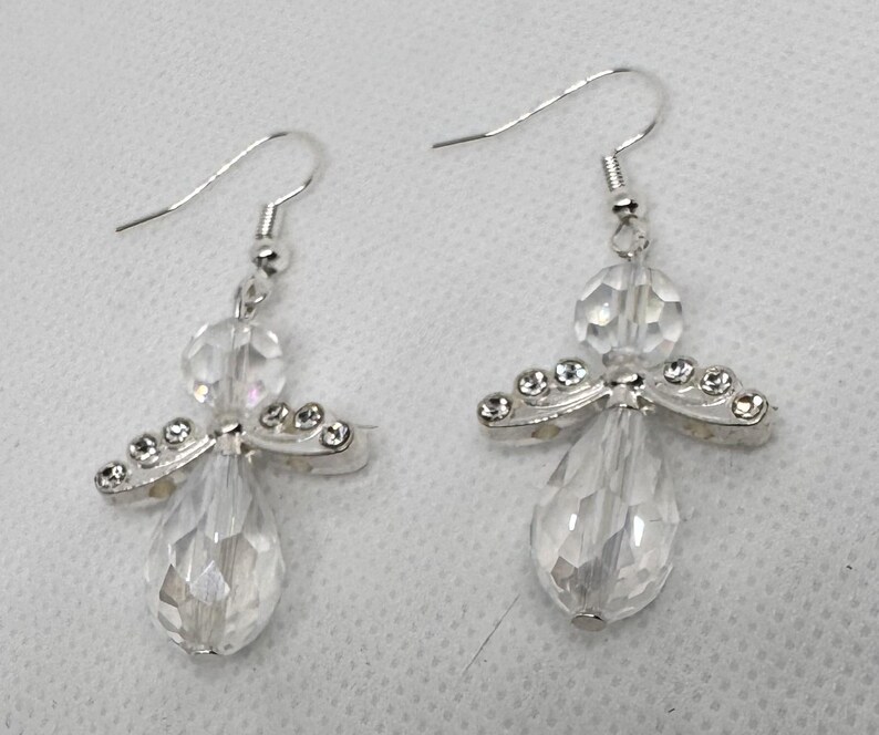May include: A pair of clear crystal angel earrings. Each earring features a teardrop-shaped crystal body, a round crystal head, and silver-toned wings adorned with small, clear crystals. The earrings have silver-colored fish hook closures.