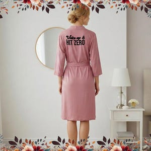 Peut inclure: Un peignoir rose poussiéreux avec le texte "Wake up & Hit Zero" imprimé au dos. La robe a une ceinture assortie et des manches trois-quarts. La personne se tient devant un mur blanc avec un miroir rond.