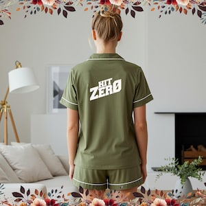 May include: An olive green pajama set with white piping, displaying the text "HIT ZERO" in white on the back. The set comprises a short-sleeved top and matching shorts. The person is standing in a bright room.