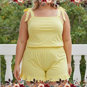 Puede incluir: Un conjunto de pijama de dos piezas de color amarillo claro. La parte superior es un top sin mangas con tirantes anudados, y los pantalones cortos tienen un dobladillo con volantes. El conjunto está hecho de una tela suave y ligera, perfecta para el clima cálido o para descansar. El fondo es una escena exterior borrosa.
