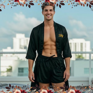 Könnte beinhalten: Ein schwarzes Set aus Satin-Bademantel und passender Shorts mit dem in Goldfaden gestickten Namen Toby auf der linken Brust. Der Bademantel hat dreiviertellange Ärmel und eine offene Vorderseite, während die Shorts einen Kordelzug besitzen.