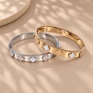 May include: Two hinged bangle bracelets, one in a polished silver tone and one in a gold tone. Each bracelet features a series of evenly spaced, four-leaf clover motifs, with some inlaid with white material and others in solid metal.