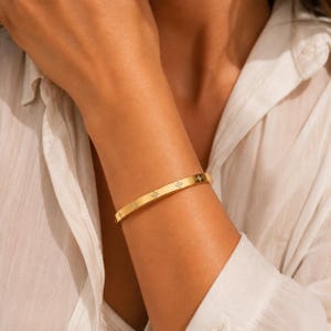 May include: A gold-toned bangle bracelet featuring a series of evenly spaced, small four-petaled flower motifs. Each flower design is accented with tiny, clear embedded stones that catch the light, creating a subtle sparkle along the polished metal band.