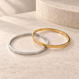 May include: Two hinged bangle bracelets, one in a polished silver tone and one in a polished gold tone. Each bracelet features a smooth, rounded band with small, evenly spaced clear stones embedded along the outer surface of the metal.