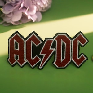 Könnte beinhalten: Gestickter Aufnäher mit dem Bandnamen "AC/DC" in roter und schwarzer Schrift, umrandet mit Weiß und einem Blitzmotiv. Im Hintergrund befinden sich ein farbenfrohes Lutscher und Blumen. Geeignet für Kleidung oder Accessoires.