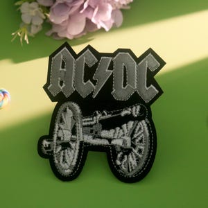 May include: Black and gray AC/DC patch featuring the band's logo and a cannon design. The patch is on a green surface with two colorful lollipops and a cluster of purple flowers.