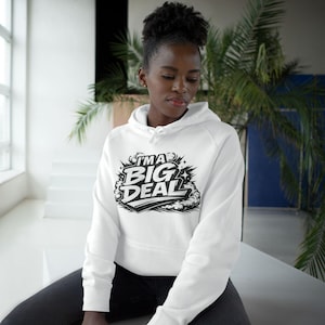 Peut inclure: Un sweat à capuche blanc avec l'inscription "I'M A BIG DEAL" en noir et blanc. Le sweat à capuche a une capuche à cordon et des manches longues. La personne porte un jean gris foncé.