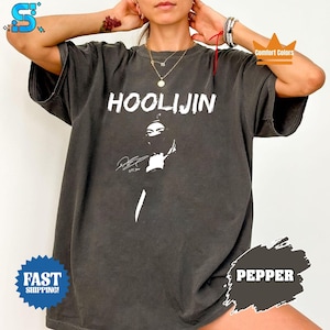 May include: Dark gray t-shirt with the word "HOOLIJIN" in white block letters. The shirt features a graphic of a person with a black face covering and a signature. The shirt is a Comfort Colors brand.