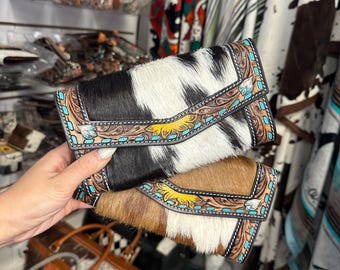 May include: Two cowhide leather wallets with turquoise and brown decorative trim. One wallet is black and white, the other is brown and white. The wallets have a flap closure and are held in front of a store display.