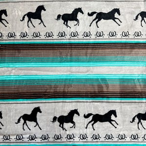 May include: A blanket with a repeating pattern of black horse silhouettes on a light gray background. The design includes horizontal stripes in turquoise, brown, and gray, with a decorative element below the horses. The blanket has a soft, plush texture.
