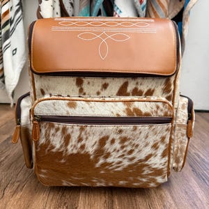 May include: A backpack featuring a brown leather top flap with white stitched decorative patterns, a main body made of brown and white cowhide, a large front zippered pocket, and two side zippered pockets with brown leather trim and dark zippers.