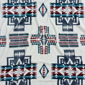 May include: A white blanket with a geometric pattern in shades of blue, burgundy, and teal. The design features repeating cross-like shapes and diamond motifs, creating a Southwestern or Native American aesthetic.