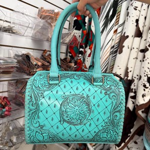 May include: A turquoise handbag featuring an intricate embossed floral pattern and a diamond-shaped grid design. The bag has two matching turquoise top handles, a zipper closure, and dark brown accents within the embossed grooves of the leather.