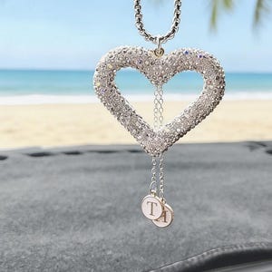 May include: A silver heart-shaped car charm, encrusted with sparkling crystals, hangs from a chain. Two small, round charms with the letter "T" dangle below. The background shows a beach and ocean.