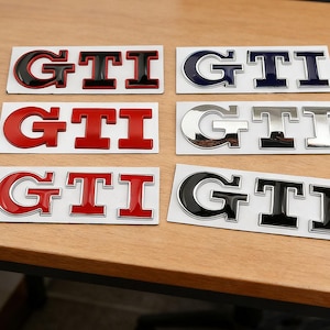 Peut inclure: Plusieurs autocollants d'emblèmes GTI de différentes couleurs, dont le rouge, le noir, l'argent et le bleu. Les autocollants sont sur un support blanc et sont présentés sur une surface en bois. Le logo GTI est bien visible.