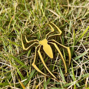 May include: A gold-colored spider-shaped decorative object with eight legs, resting on a bed of green grass. The spider has a shiny, metallic finish and is likely made of metal. The legs are long and slender, and the body is a solid, rounded shape.