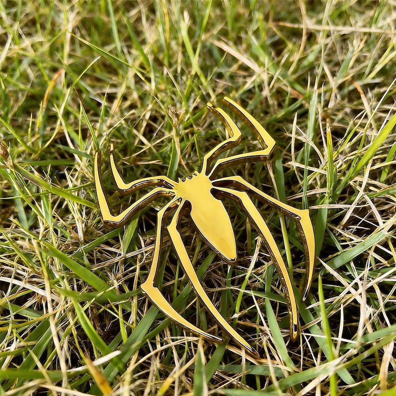 May include: A gold-colored spider-shaped decorative object with eight legs, resting on a bed of green grass. The spider has a shiny, metallic finish and is likely made of metal. The legs are long and slender, and the body is a solid, rounded shape.