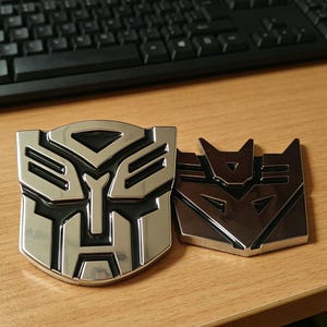 Puede incluir: Dos emblemas cromados de Transformers, uno Autobot y otro Decepticon, sobre una superficie de madera. El emblema Autobot es un escudo plateado con contorno negro. El emblema Decepticon es una forma angular plateada.