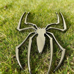 May include: A shiny, silver-colored spider emblem with eight legs, resting on a bed of green grass. The spider's body is a teardrop shape, and the legs curve outwards. The emblem is likely a decorative item or a car emblem.