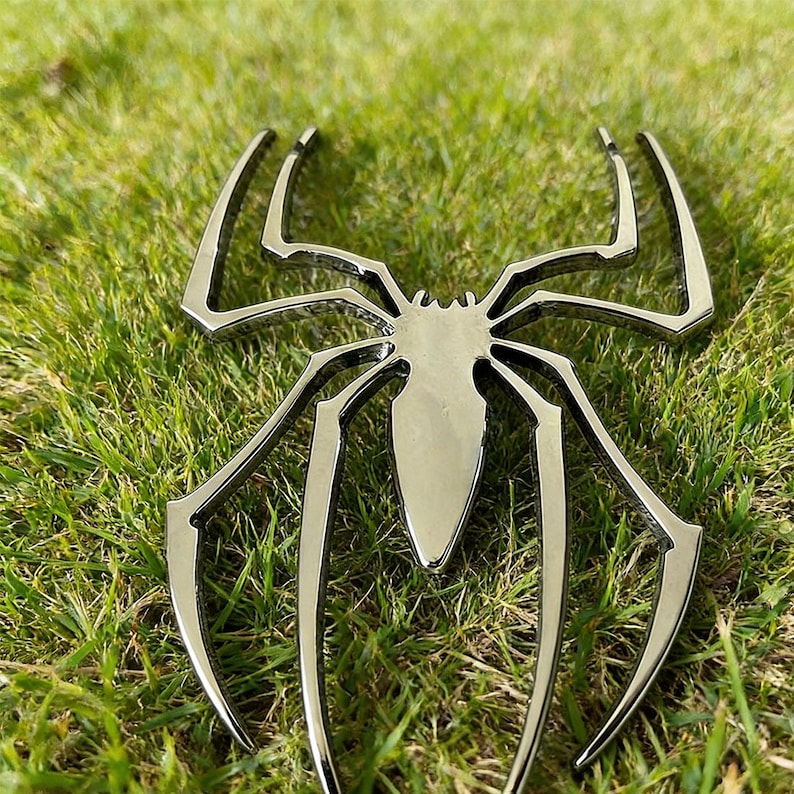 May include: A shiny, silver-colored spider emblem with eight legs, resting on a bed of green grass. The spider's body is a teardrop shape, and the legs curve outwards. The emblem is likely a decorative item or a car emblem.