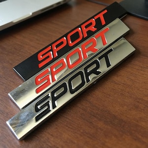 May include: Three rectangular car emblems with the word "SPORT" in bold, capital letters. One is black with red lettering, and two are silver with red and black lettering. The emblems are arranged diagonally on a wooden surface.