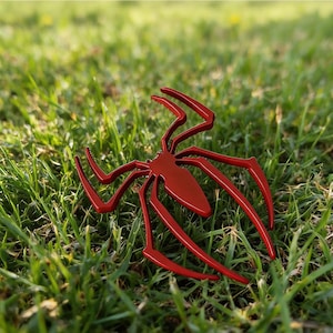 May include: A glossy, red spider emblem with long, curved legs rests on a bed of green grass. The spider's design is sleek and modern, with a smooth, reflective surface. The emblem is a striking decorative element.