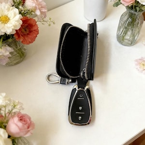 May include: Black leather key case with a silver zip and a silver key ring. The case is open, revealing the interior. A car key is attached. Flowers in vases are in the background.