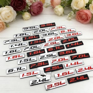 Puede incluir: Una colección de emblemas de coche plateados y negros con letras rojas, que muestran tamaños de motor como 1.5L, 2.0L y 3.5L. Los emblemas están dispuestos sobre una superficie blanca, con flores en el fondo.