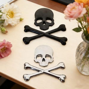May include: Two skull and crossbones decorations. One is black, the other is silver. The crossbones are black and silver. The decorations are on a light wood surface. Flowers in a vase are in the background.