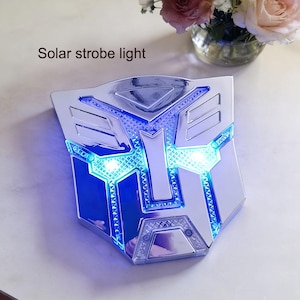 May include: A silver solar strobe light shaped like a stylized emblem, with blue and white LED lights. The text "Solar strobe light" is above the emblem. The light is on a white surface.