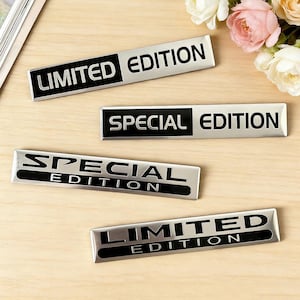May include: Four rectangular metal badges with the words "LIMITED EDITION" and "SPECIAL EDITION" in black lettering. The badges have a silver finish and are arranged on a light wood surface. A bouquet of flowers is in the background.