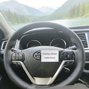 Puede incluir: Volante de cuero negro con detalles perforados y detalles plateados. El volante tiene controles integrados y un emblema central de Toyota. Una pegatina blanca dice "Overlay Sticker matte black".