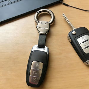 May include: A black car key fob with silver detailing and a key ring featuring an Iron Man mask design. The key ring is attached to a black leather strap. Another car key fob is visible on a wooden surface.