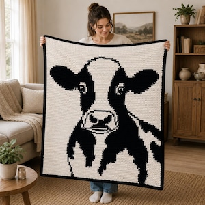 May include: A rectangular crochet blanket featuring a large, centered graphic of a cow's face in high-contrast black and white yarn. The design displays the cow's ears, eyes, and snout in a pixelated style against a solid off-white background.