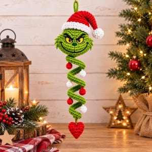May include: A crocheted hanging ornament featuring a green character head with a mischievous expression, wearing a red and white Santa hat. Below the head is a green spiral tail decorated with small red and white spheres, ending in a red heart.