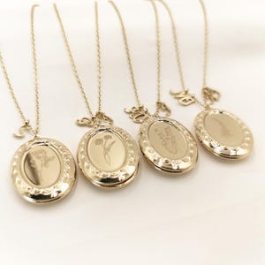 May include: Four gold-colored oval lockets on delicate chains. Each locket features a floral engraving and a decorative border. One locket has a letter charm. The lockets are displayed on a white surface.