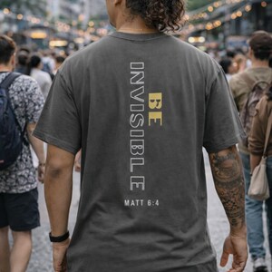May include: Dark gray t-shirt with the words "BE INVISIBLE" printed vertically in white and gold. The text "MATT 6:4" is printed below. The shirt is worn by a person walking in a crowded outdoor setting.