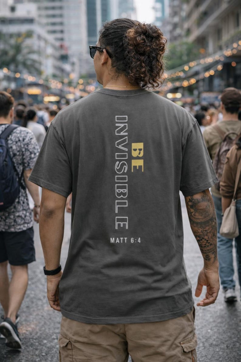 May include: Dark gray t-shirt with the words "BE INVISIBLE" printed vertically in white and gold. The text "MATT 6:4" is printed below. The shirt is worn by a person walking in a crowded outdoor setting.