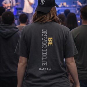 May include: A person wearing a black baseball cap with a white "NY" logo and a gray t-shirt. The t-shirt has the words "BE INVISIBLE" and "MATT 6:4" printed on the back. The person is in a crowd.