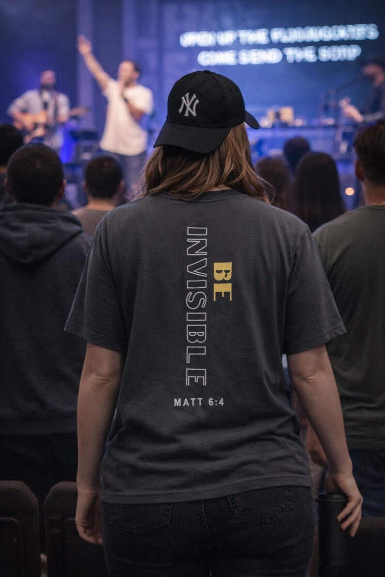 May include: A person wearing a black baseball cap with a white "NY" logo and a gray t-shirt. The t-shirt has the words "BE INVISIBLE" and "MATT 6:4" printed on the back. The person is in a crowd.
